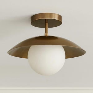 Può includere: Una plafoniera in ottone con un paralume bianco a forma di globo. L'apparecchio ha una base circolare e un paralume ampio e curvo. Il design è moderno e minimalista, adatto a vari stili d'interni.