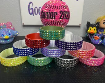 May include: A collection of colorful, bedazzled bracelets in various colors, including red, green, blue, purple, and pink. The bracelets are stacked, showcasing their sparkling rhinestone embellishments. A pink bedazzled softball and a sign that says "Good Senior 2023" are in the background.