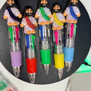 May include: Five multi-color ink pens featuring a decorative topper of a figure with long dark hair and a beard, wearing a white robe covered in iridescent rhinestones with a diagonal sash in purple, red, green, yellow, or blue.