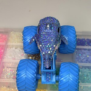 Op de afbeelding: Een blauw monstertruckspeelgoed met grote, getextureerde banden. De carrosserie van de truck heeft de vorm van een dolfijn en is bedekt met iriserende strass-steentjes. Het speelgoed staat tegen een achtergrond van kleine, kleurrijke knutselspullen.