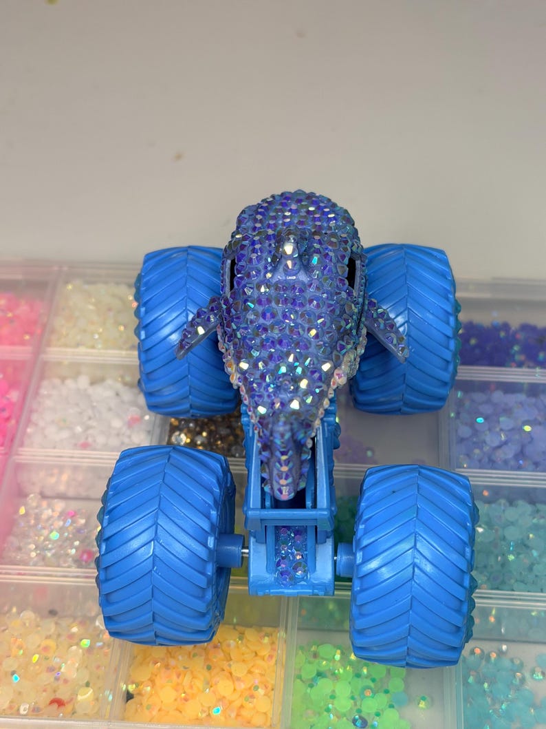 Op de afbeelding: Een blauw monstertruckspeelgoed met grote, getextureerde banden. De carrosserie van de truck heeft de vorm van een dolfijn en is bedekt met iriserende strass-steentjes. Het speelgoed staat tegen een achtergrond van kleine, kleurrijke knutselspullen.