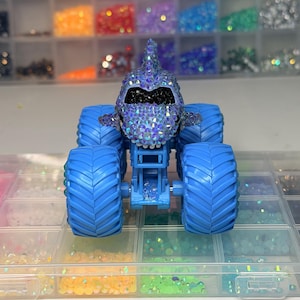 Op de afbeelding: Een blauw monstertruckspeelgoed met een haai-vormig lichaam bedekt met iriserende strass-steentjes. De truck heeft grote blauwe banden en staat tegen een achtergrond van kleurrijke knutselspullen, wat wijst op een op maat gemaakt of handgemaakt item.
