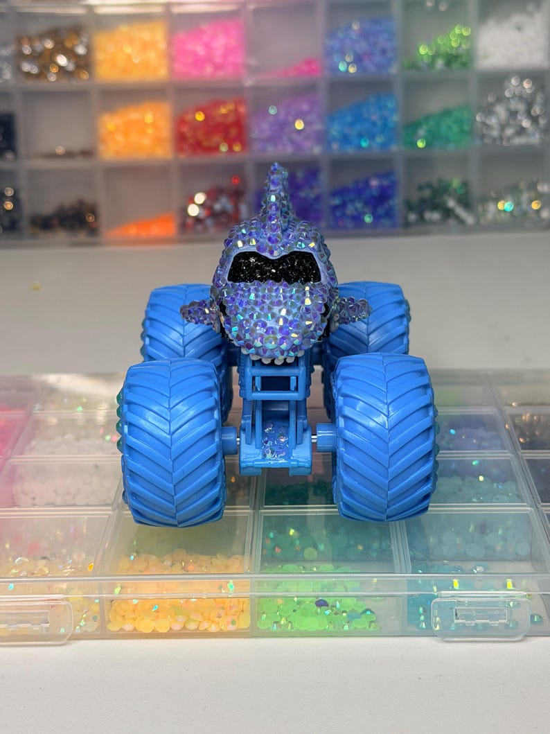Op de afbeelding: Een blauw monstertruckspeelgoed met een haai-vormig lichaam bedekt met iriserende strass-steentjes. De truck heeft grote blauwe banden en staat tegen een achtergrond van kleurrijke knutselspullen, wat wijst op een op maat gemaakt of handgemaakt item.