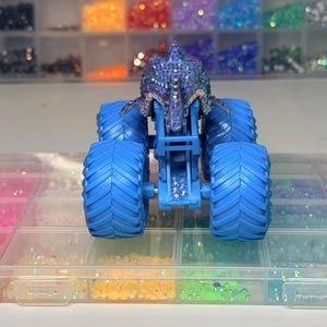 Op de afbeelding: Een blauwe monstertruck speelgoed met grote, getextureerde banden. De carrosserie van de truck is versierd met iriserende strass-steentjes. De achtergrond toont bakjes gevuld met kleurrijke strass-steentjes, wat wijst op een knutsel- of hobbymotief.
