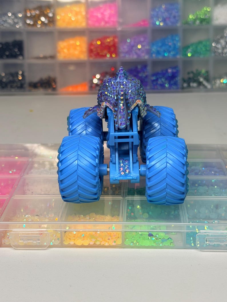 Op de afbeelding: Een blauwe monstertruck speelgoed met grote, getextureerde banden. De carrosserie van de truck is versierd met iriserende strass-steentjes. De achtergrond toont bakjes gevuld met kleurrijke strass-steentjes, wat wijst op een knutsel- of hobbymotief.