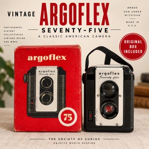 May include: A black Argoflex Seventy-five camera with a silver front plate, dual lenses, and a black carrying strap, positioned next to its original red cardboard box featuring the camera name, a graphic illustration, and the number 75.