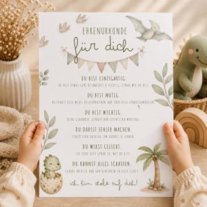 May include: A white certificate with the German text "EHRENURKUNDE für dich" featuring watercolour illustrations of a pterodactyl, a baby dinosaur in an egg, a palm tree, and green leaves. The document includes positive affirmations in German.