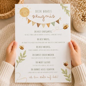 May include: A white paper print titled "DEIN WAHRES ZEUGNIS" featuring a yellow sun, colourful bunting, and bee illustrations. The text includes affirmations like "DU BIST EINZIGARTIG" and "DU BIST MUTIG" written in a dark brown script and print font.