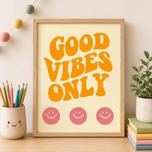 Könnte beinhalten: Ein gerahmter Kunstdruck mit dem orangefarbenen Text GOOD VIBES ONLY in einer wellenförmigen, retro-inspirierten Schrift auf hellcremefarbenem Hintergrund, ergänzt durch drei rosa Smiley-Symbole in einer horizontalen Reihe am unteren Rand.