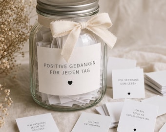 May include: A clear glass jar with a silver lid, filled with small white paper slips. A cream-colored ribbon is tied around the neck. The label reads "POSITIVE GEDANKEN FÜR JEDEN TAG" with a heart icon. Several loose paper slips with German text and heart icons are scattered nearby.