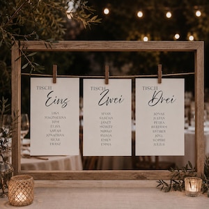 May include: A wooden framed seating plan with three white cards suspended from a string. Each card displays a table number in German and a list of names. The frame has a rustic appearance, with soft lighting in the background.