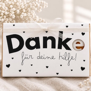 Könnte beinhalten: Eine rechteckige weiße Geschenkbox mit dem schwarzen Schriftzug Danke für deine hilfe! und kleinen schwarzen Herzsymbolen. Die Box ist mit einem dünnen weißen Band verschlossen und hat einen runden Ausschnitt, der den Inhalt zeigt.