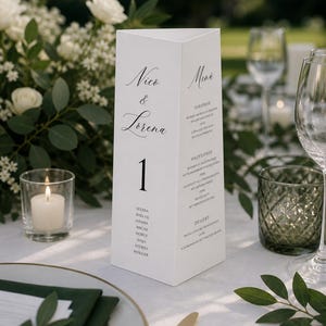 May include: A white, triangular table card with the names "Nico & Lorena" in elegant script. The number "1" is prominently displayed below. Additional text includes a menu and guest names. The card is set on a white tablecloth.