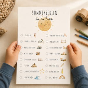 Op de afbeelding: Een witte papieren checklist getiteld SOMMERFERIEN to do liste met een centrale gele zonne-illustratie. Het bevat twee kolommen met Duitse tekstitems zoals Eis essen, Fahrrad fahren en Sterne beobachten, elk vergezeld van een klein icoontje.
