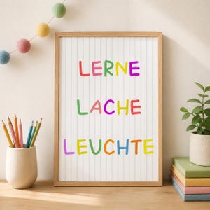 Op de afbeelding: Een ingelijste verticale wandprint met de Duitse woorden LERNE, LACHE en LEUCHTE in kleurrijke, handgetekende letters tegen een witte achtergrond met dunne verticale grijze strepen, weergegeven in een lichthouten lijst.