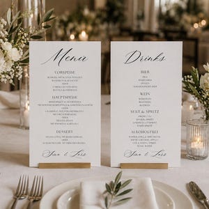 Peut inclure: Mise en place élégante pour un mariage, avec deux menus et des compositions florales. Les cartes, intitulées "Menu" et "Boissons", sont posées sur des supports en bois. Roses blanches et bougies créent une ambiance romantique. Verres à vin et couverts.
