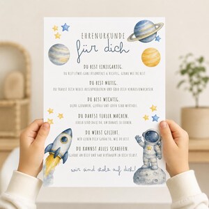 Op de afbeelding: Een wit certificaat getiteld Ehrenurkunde für dich met aquarelillustraties van planeten, sterren, een raket en een astronaut op de maan. De tekst bevat Duitse affirmaties over uniek zijn, moedig zijn en geliefd zijn door anderen.