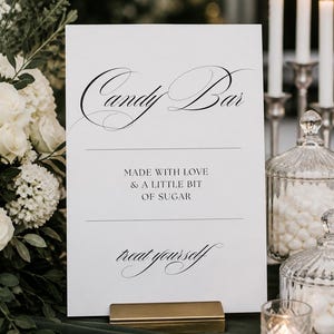 May include: A white sign with the words "Candy Bar" in elegant script. The sign also reads "Made with love & a little bit of sugar" and "treat yourself." The sign is held by a gold-coloured metal holder. The background includes white flowers, candles, and glass jars.