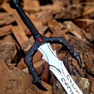 May include: A fantasy sword with a silver blade and black handle. The handle features a red gem and intricate designs. The sword's hilt has a unique claw-like shape. The blade has an inscription.