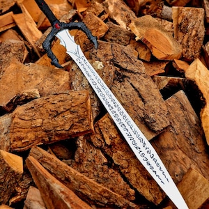 May include: A fantasy sword with a black handle and guard, featuring a red gem. The silver blade is inscribed with an unknown script. The sword rests on a pile of brown firewood.