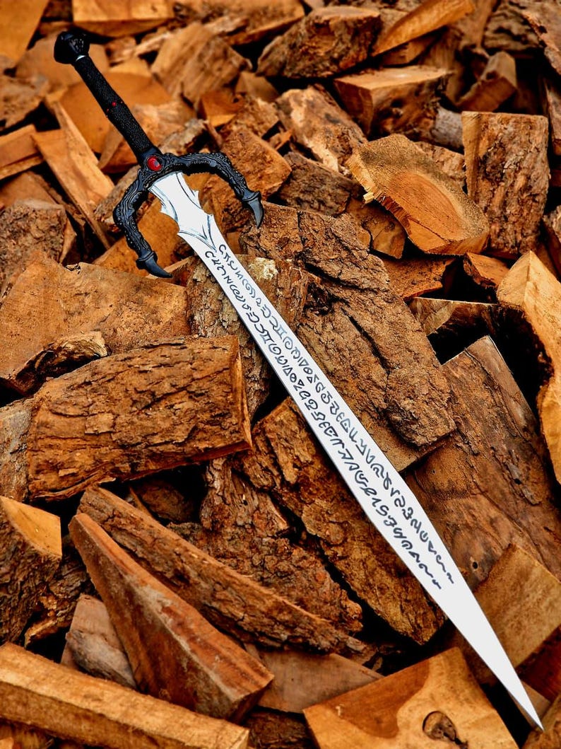 May include: A fantasy sword with a black handle and guard, featuring a red gem. The silver blade is inscribed with an unknown script. The sword rests on a pile of brown firewood.