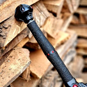 May include: A black fantasy sword with a skull-shaped pommel and red gem accents. The handle has a textured grip, and the blade guard features ornate designs and a central red gem. The sword is set against a backdrop of stacked firewood.