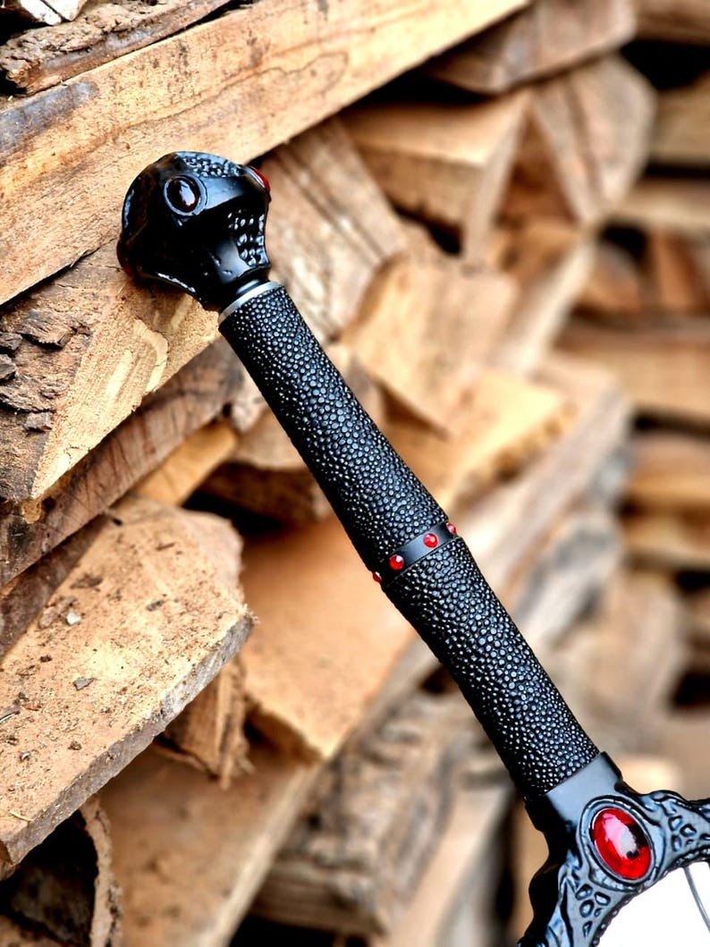 May include: A black fantasy sword with a skull-shaped pommel and red gem accents. The handle has a textured grip, and the blade guard features ornate designs and a central red gem. The sword is set against a backdrop of stacked firewood.