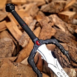 May include: A detailed fantasy sword with a black textured handle and guard, featuring a red gem and intricate designs. The silver blade has etched inscriptions. The sword is displayed against a backdrop of wooden blocks.