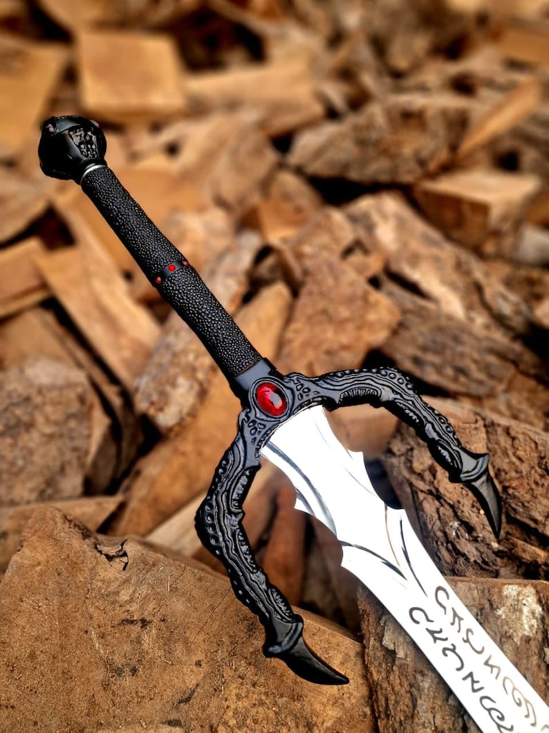 May include: A detailed fantasy sword with a black textured handle and guard, featuring a red gem and intricate designs. The silver blade has etched inscriptions. The sword is displayed against a backdrop of wooden blocks.