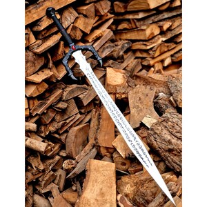May include: A long, silver sword with a black handle and guard, featuring a red gem and intricate designs. The blade has etched text. The sword is displayed against a backdrop of stacked firewood.