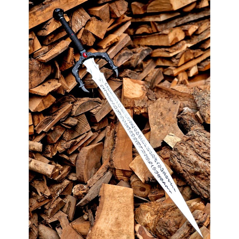 May include: A long, silver sword with a black handle and guard, featuring a red gem and intricate designs. The blade has etched text. The sword is displayed against a backdrop of stacked firewood.