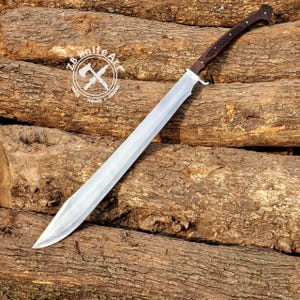May include: A large, silver-bladed sword with a dark wood handle and a decorative hilt. The blade is long and straight, with a sharp point. The handle features a textured grip. The sword rests on a background of stacked logs, with a logo in the upper left corner.