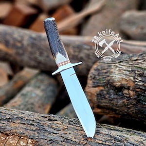 Puede incluir: Un cuchillo de caza artesanal con una hoja de acero pulido y un mango de asta texturizado. El cuchillo presenta una guarda y un anillo decorativo cerca del mango. El cuchillo se exhibe sobre una superficie de madera.