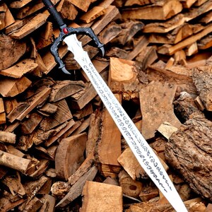 May include: A decorative sword with a black handle and guard, featuring a red gem and ornate detailing. The silver blade has etched inscriptions. The sword rests against a backdrop of stacked firewood, creating a rustic aesthetic.