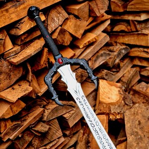 May include: A detailed fantasy sword with a black handle and guard, featuring a red gem and intricate designs. The silver blade has engraved text. The sword is set against a backdrop of stacked firewood.