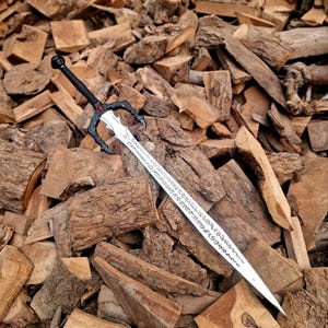 May include: A silver sword with a black handle and a decorative guard, resting on a pile of wood. The blade has etched inscriptions. The handle has a red gem. The sword is a fantasy-themed item.