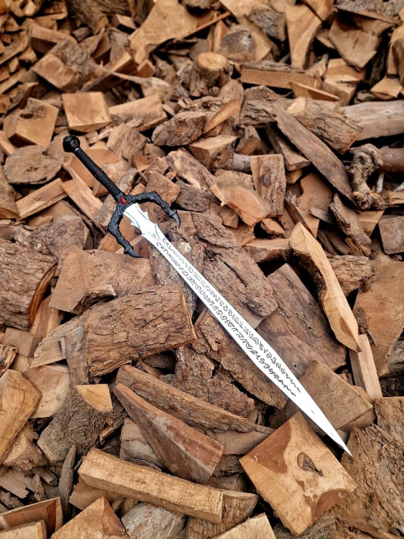 May include: A silver sword with a black handle and a decorative guard, resting on a pile of wood. The blade has etched inscriptions. The handle has a red gem. The sword is a fantasy-themed item.