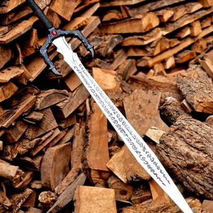 May include: A decorative sword with a black handle and a silver blade. The handle features a red gem and ornate detailing. The blade has etched inscriptions. The sword is displayed against a backdrop of stacked firewood.