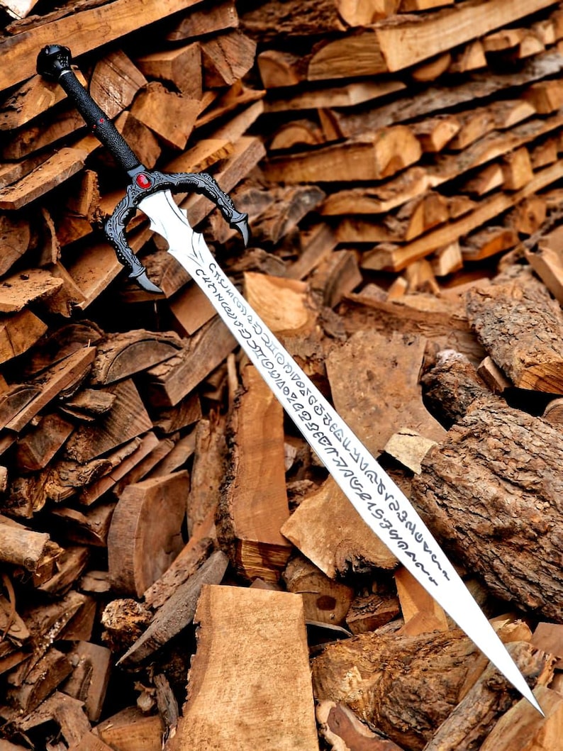 May include: A decorative sword with a black handle and a silver blade. The handle features a red gem and ornate detailing. The blade has etched inscriptions. The sword is displayed against a backdrop of stacked firewood.