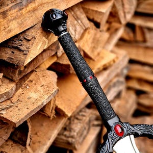 May include: A black fantasy sword with a textured grip and ornate details. The hilt features a skull design and red accents. The blade is silver and the sword is resting on a pile of wood.