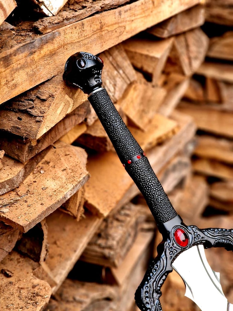 May include: A black fantasy sword with a textured grip and ornate details. The hilt features a skull design and red accents. The blade is silver and the sword is resting on a pile of wood.