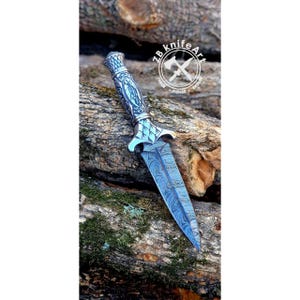 May include: A decorative dagger with a patterned blue blade and ornate silver handle. The handle features detailed carvings and a textured grip. The blade has a unique wave-like pattern. The knife is displayed on a wooden surface, with the text "ZB knife Art".