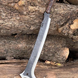 May include: A long, heavy-duty machete with a dark, textured forged steel blade featuring a sharpened edge, a curved tip with a circular cutout, and three small holes near the handle. The handle is made of dark brown wood with three brass rivets.