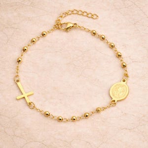 Könnte beinhalten: Goldfarbenes Armband mit einem Kreuzanhänger und einem runden Anhänger mit detailliertem Relief. Das Armband besteht aus kleinen, runden Perlen, die durch eine filigrane Kette verbunden sind. Der Verschluss ist am Ende der Kette sichtbar.