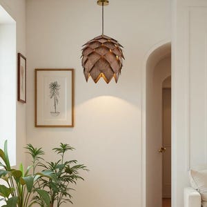 May include: A decorative pendant light fixture with a layered, brown, artichoke-like design hangs from the ceiling. The light fixture is suspended in a hallway with white walls, a wooden floor, and potted plants. A framed botanical print hangs on the wall.