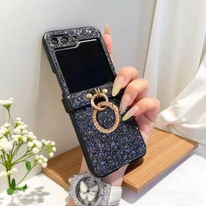 May include: A black glitter phone case with a gold ring holder. The case has a rectangular shape with a camera cutout and a flip-up design. The ring holder is adorned with small, sparkling crystals.