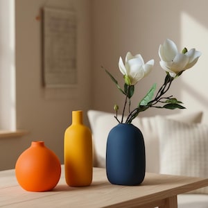 May include: Three ceramic vases in orange, yellow, and navy blue sit on a wooden table. The navy vase holds white magnolia flowers. The vases have a matte finish and a minimalist design, suitable for home decor.