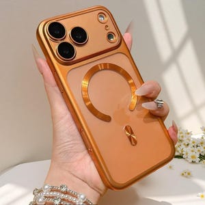 May include: A metallic gold phone case with a clear back, featuring a circular magnetic design. The case has a raised camera bezel and a small, gold accent at the bottom. The phone case is designed to protect the phone.