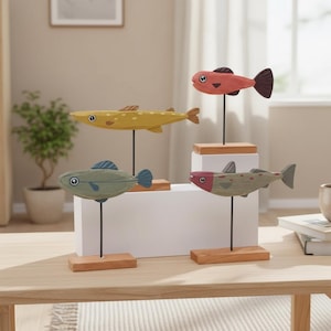 May include: Four wooden fish sculptures in shades of yellow, green, pink, and gray, each mounted on a black rod and a wooden base. The fish have painted details and are arranged on a white block and a wooden table, creating a decorative display.