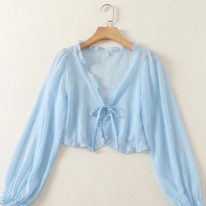 May include: A light blue, sheer bolero top with long sleeves and a tie closure. The bolero has a ruffled trim along the neckline and hem. The top is hanging on a wooden hanger against a plain background.